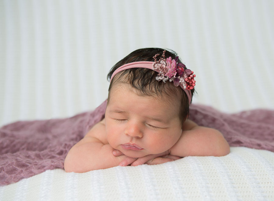 10 day old newborn baby girl laying on a cream backdrop with her tummy with her head on her hands with a pink wrap and pink headband on in a photography studio