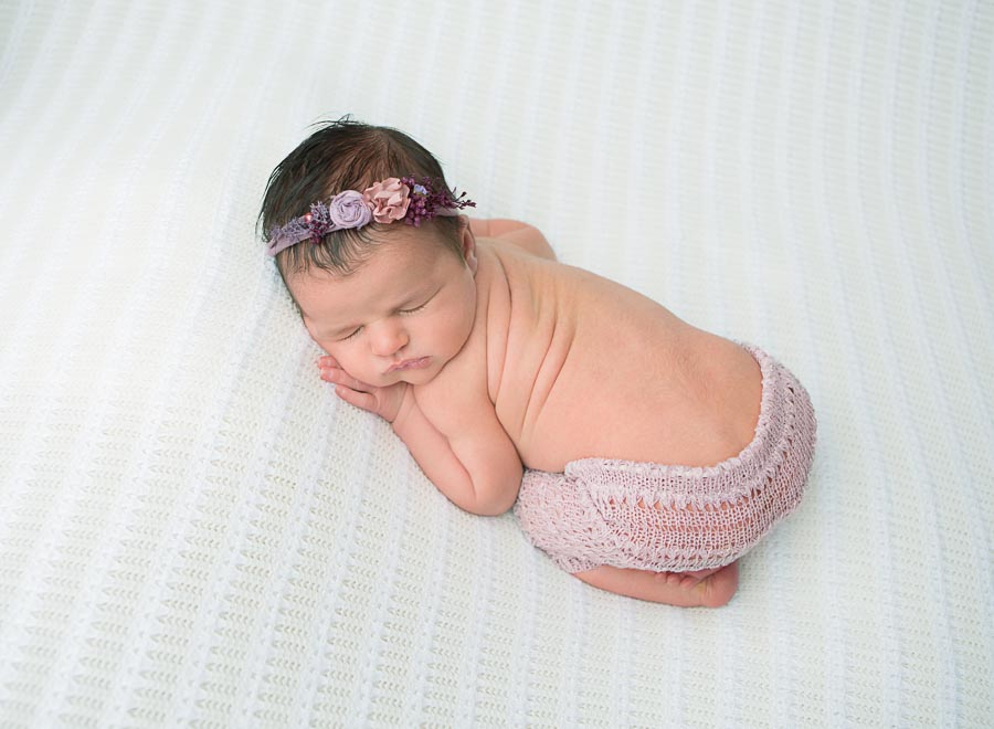 10 day old newborn baby girl posed in purple pants and purple headband posed on a cream backdrop in a photography studio
