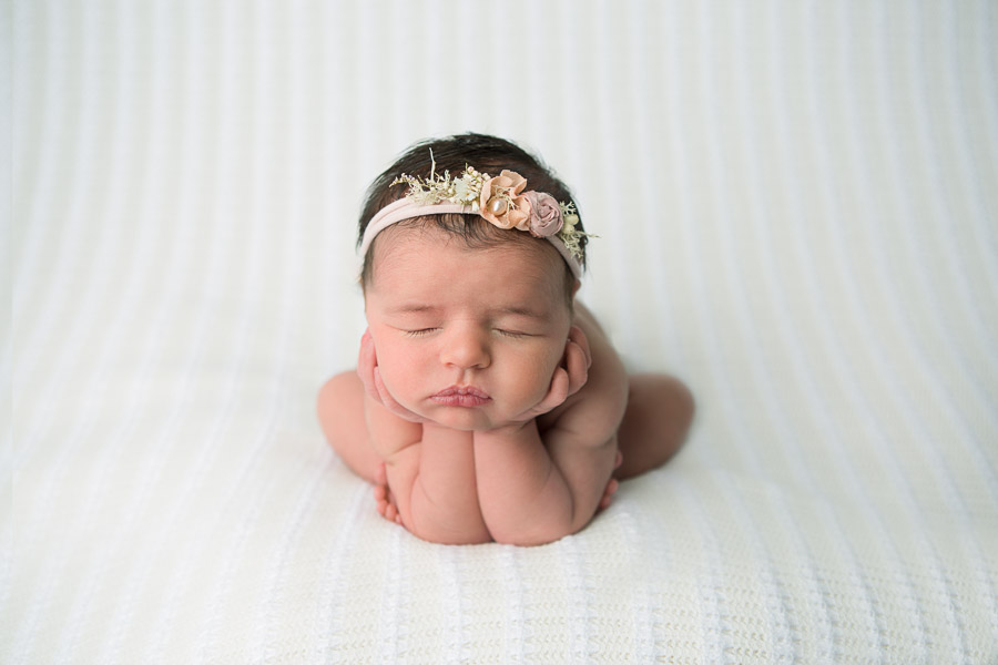 10 day old newborn baby girl posed in the frog pose on a cream backdrop with a pink headband with flowers in a photography studio