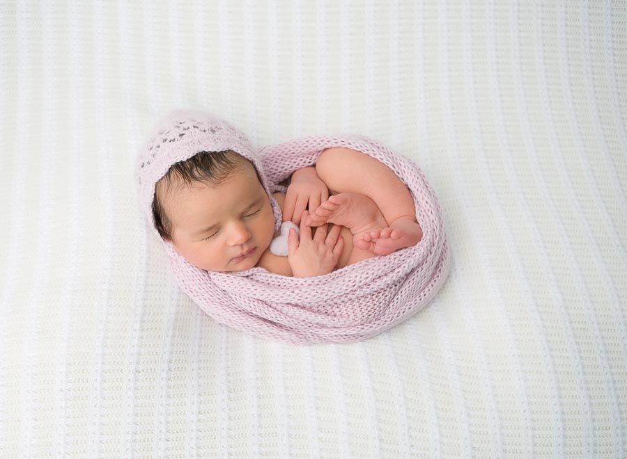 a 10 day old newborn baby girl posed on a cream backdrop wrapped in a pink wrap and pink bonnet holding a little pink heart in a photography studio