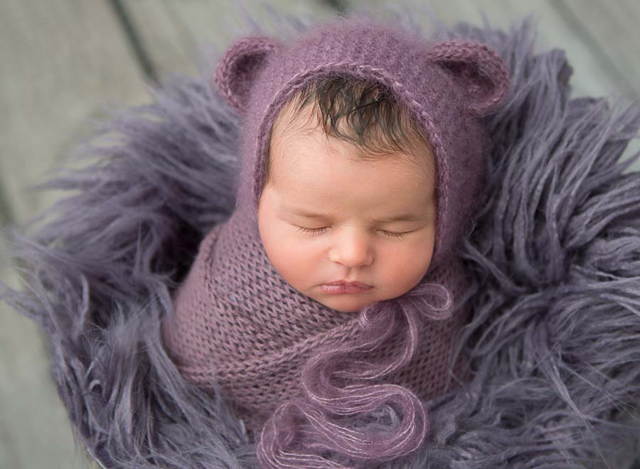 10 day old baby girl in a purple wrap and purple bonnet posed in a bucket with a purple sag material in a photography studio