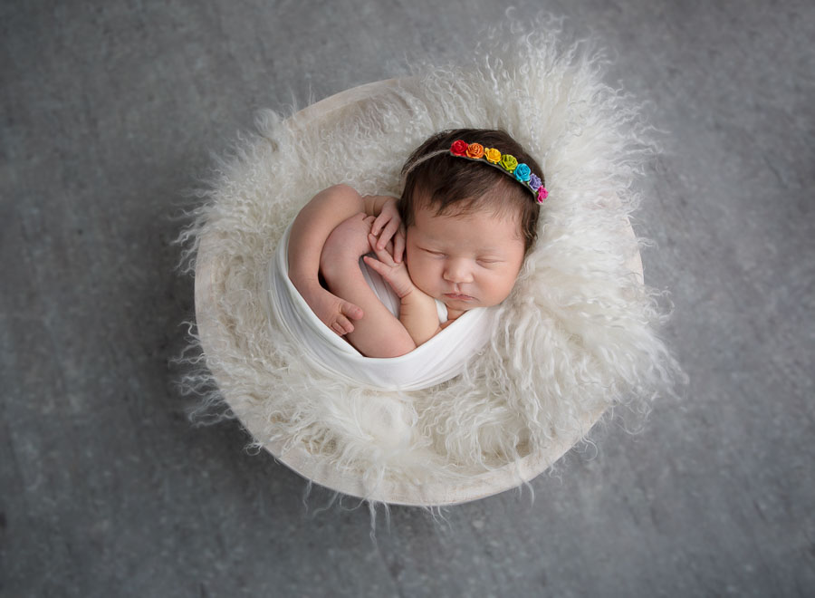 10 day old newborn baby girl wrapped in a white wrap and with a rainbow headband posed in a cream bowl on a grey backdrop in a photography studio