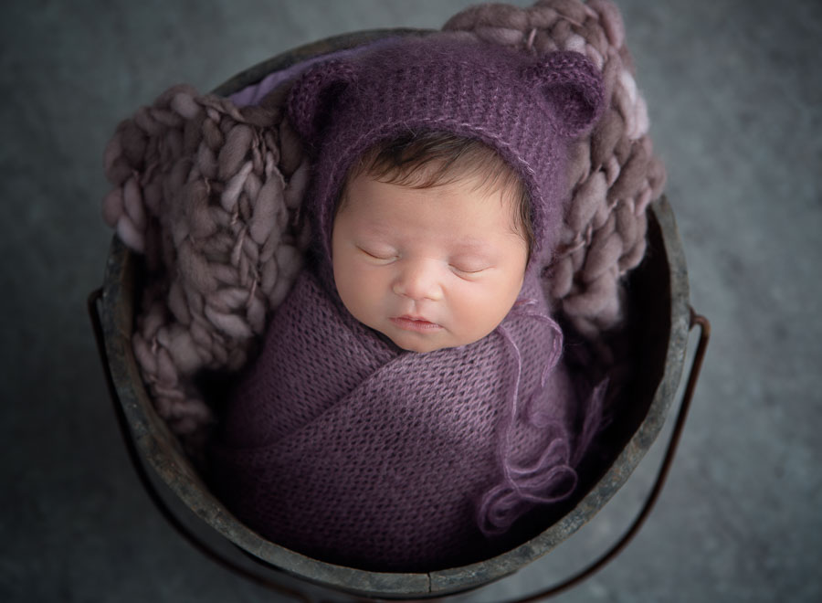 newborn baby girl 9 days old in a purple wrap and purple teddy bear bonnet posed in a bucket in a photography studio