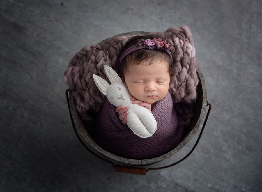 9 day old newborn baby girl in a purple wrap and headband posed in a bucket holding a bunny in a photography studio