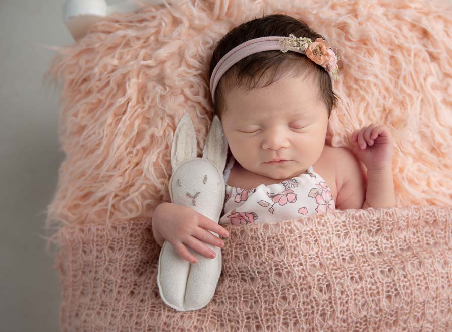 10 day old newborn baby girl with a headband on laying in a baby bed holding a little stuffed bunny