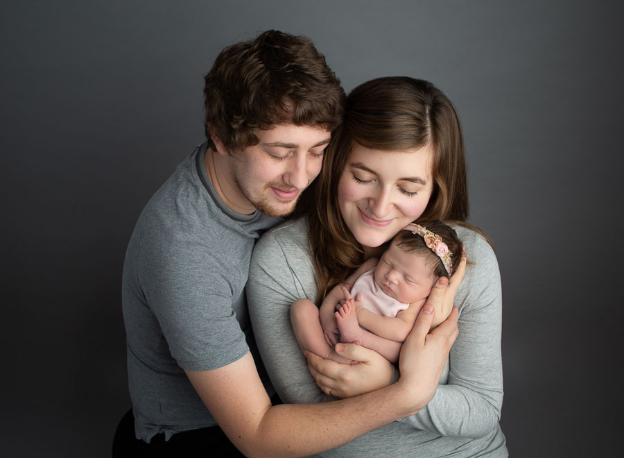 mum holding newborn baby girl with dad's arms around both of them in a photography studio.