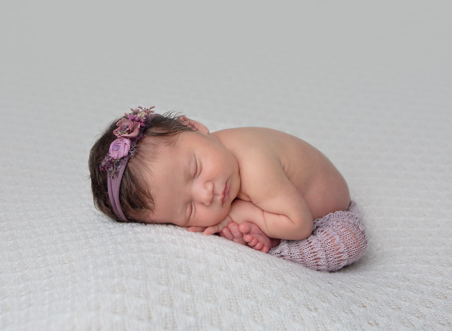10 day old newborn baby girl in purple pants and purple headband pose in the womb pose on a cream backdrop in a photography studio
