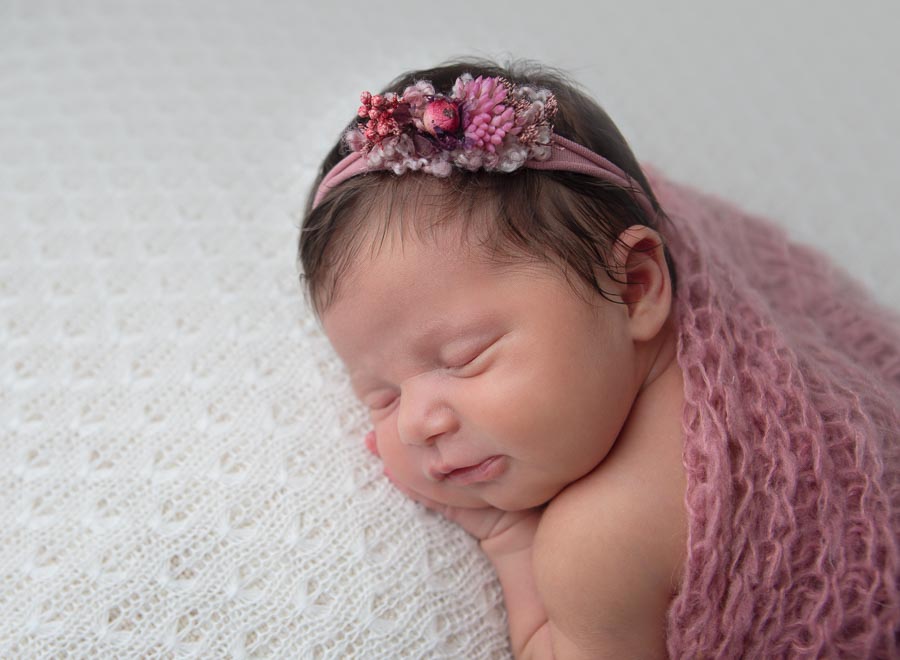 newborn baby girl 10 days old with a pink wrap and headband on posed on her tummy on a cream backdrop in a photography studio