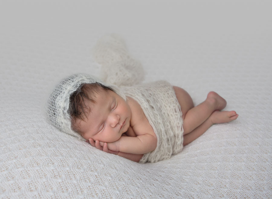 newborn baby girl in a cream wrap and bonnet posed on her side with her hands under her cheek laying on a cream backdrop in a photography studio