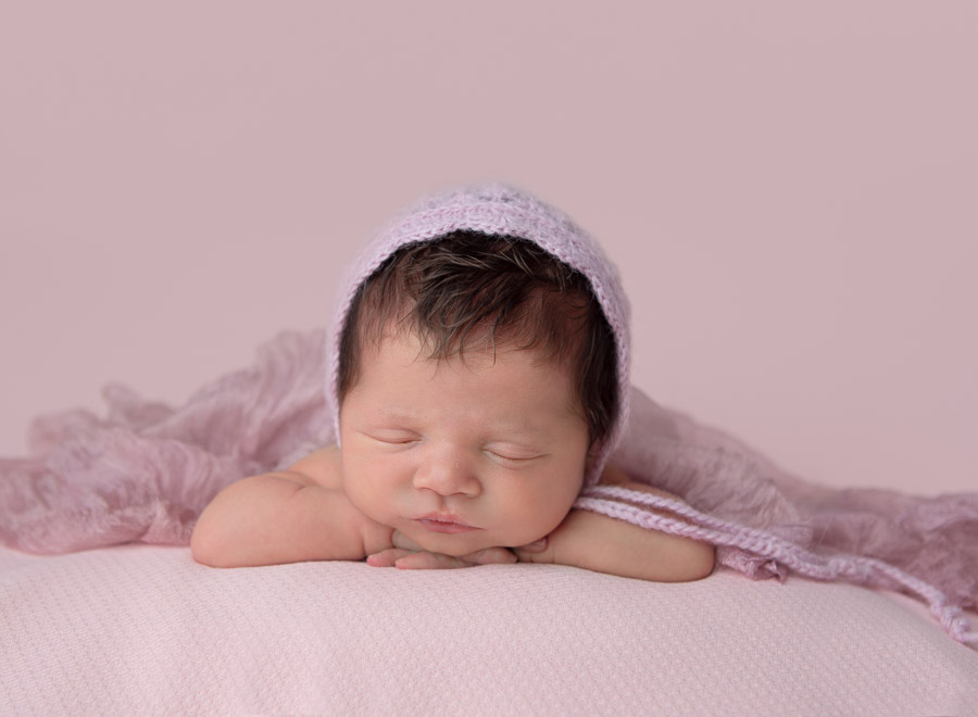 newborn baby girl at 10 days old posed with head on her hands with a pink bonnet and wrap on in a photography studio