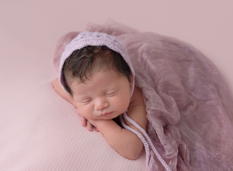 newborn baby girl at 10 days old posed with head on her hands with a pink bonnet and wrap on in a photography studio