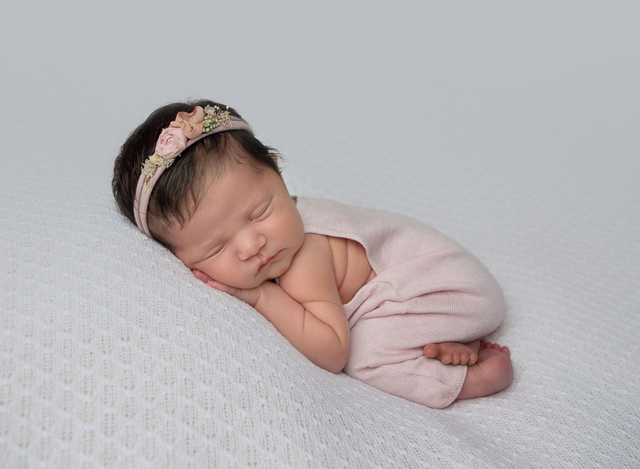 10 day old newborn baby girl with a pink outfit and headband on posed on a cream backdrop in a photography studio