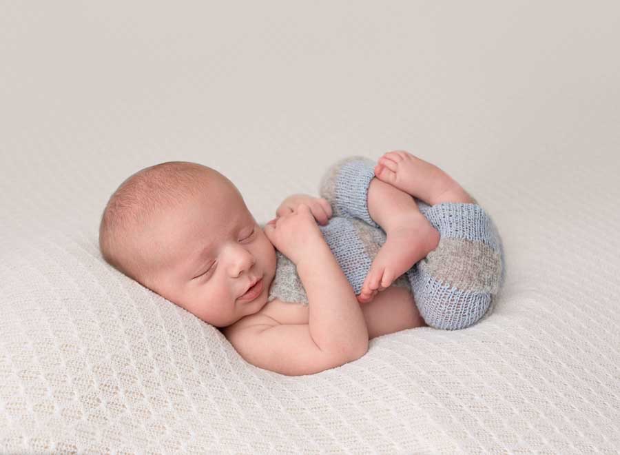 Newborn baby boy posed on his back with his legs curled up and his hand on his chest wearing a knitting blue and grey outfit laying on a cream backdrop in a studio in Doncaster