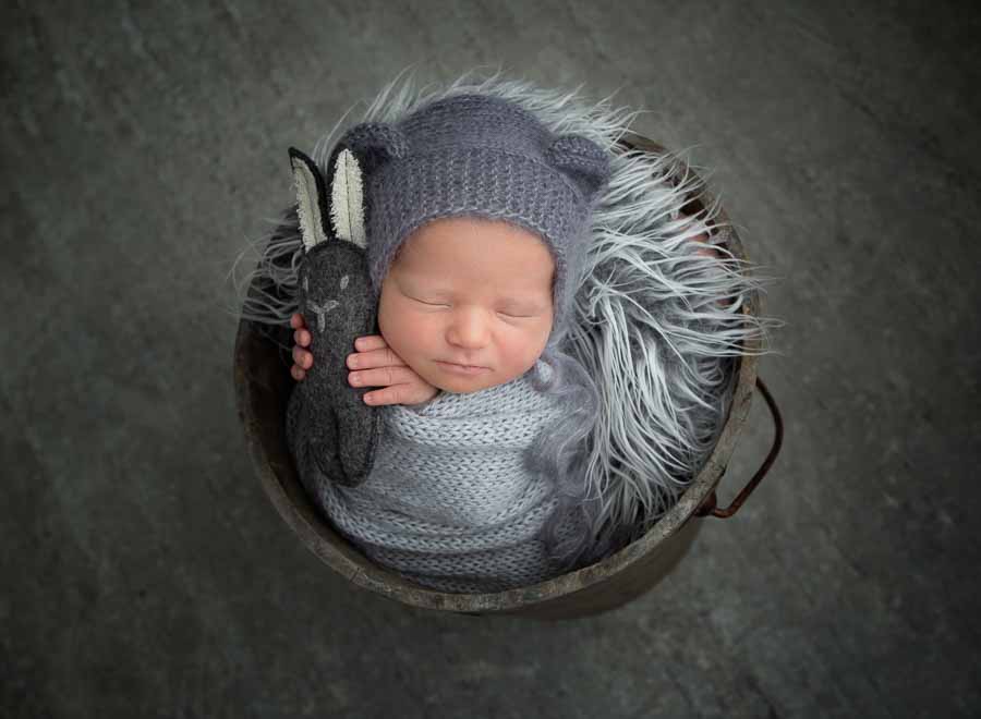Kaydee's Newborn Baby boy in a bucket with grey fur wearing a grey bonnet, holding a grey bunny on a grey studio backdrop in Doncaster
