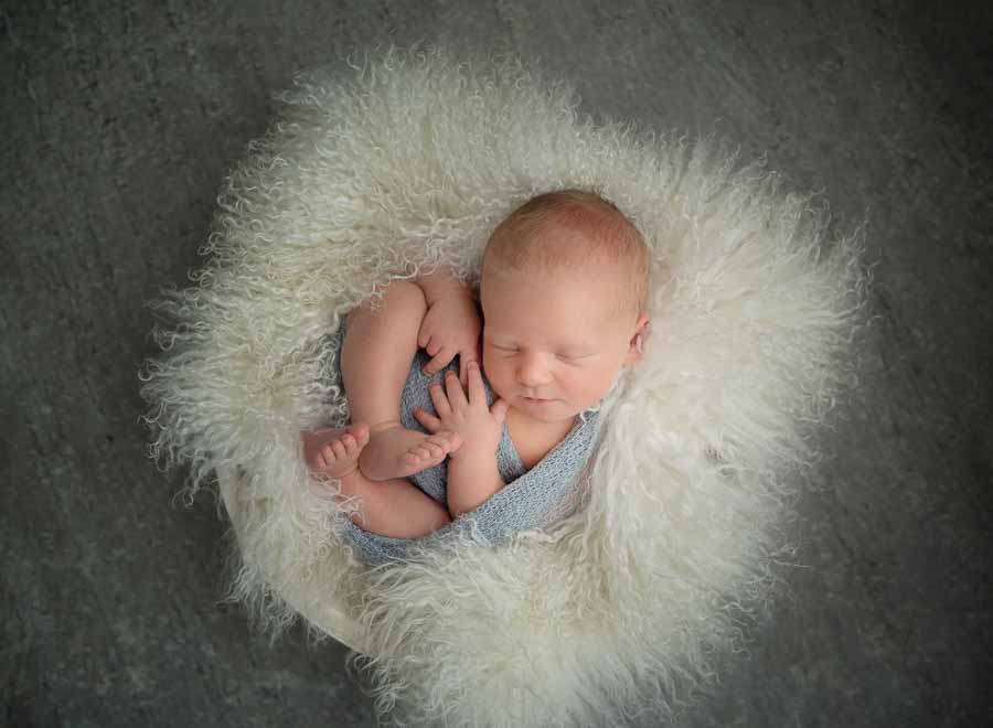 Kaydee's Newborn baby boy wrapped in a blue wrap posed on cream fur in a cream bowl on a grey wooden floor in a photography studio in Doncaster
