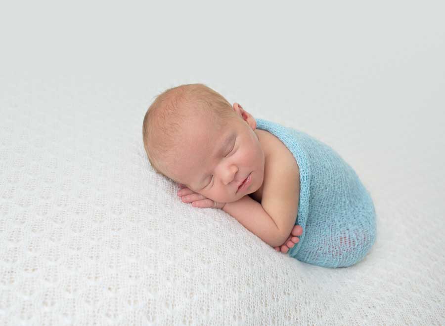 9 day old baby boy posed in the taco pose wrapped in a blue wrap on a cream backdrop in a photography studio