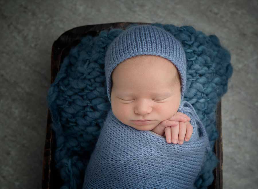 Kaydee's 10 day old Newborn Boy in a wooden dough bowl laying on a blue knitted blanket wrapped in a blue wrap with a blue bonnet with his hands crossed and poking out of the top next to his cheek.