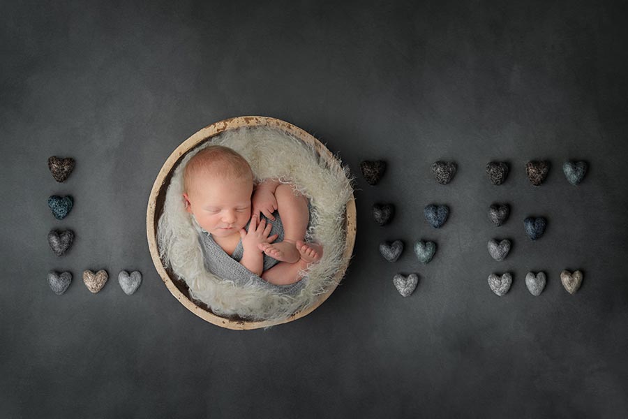 Kaydee's Ten day old newborn baby boy wrapped in a blue wrap in a wooden bowl that makes up the letter O in LOVE. The LVE are in grey and blue felt hearts