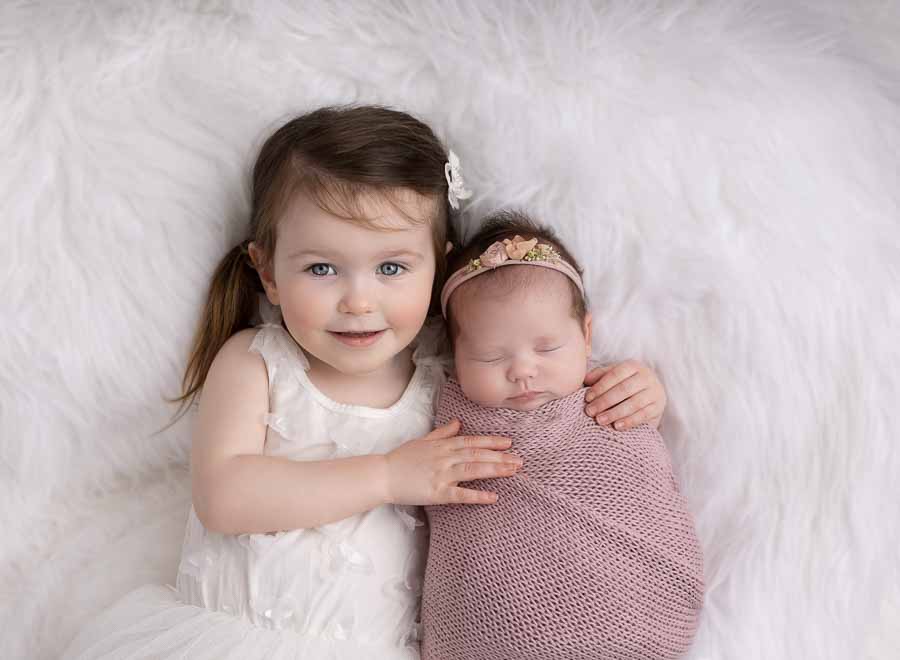Big sister is holding her new baby sister in her arms on a fluffy white rug during a newborn session in Doncastser.